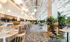 Moderni ja valoisa ravintola, jossa on pöytiä ja tuoleja sekä vihreitä kasveja. Korkeat katot ja valaistus luovat ilmavan tunnelman. (Tekoälyn luoma teksti)