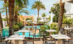 Kuva ravintolaterassista, josta on näkymä uima-altaalle, jota ympäröivät palmut ja vaaleat rakennukset. (Tekoälyn luoma teksti)