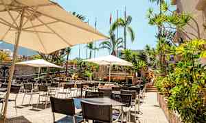 Kuva terassilta, jossa on pöytiä, tuoleja ja aurinkovarjoja trooppisessa ympäristössä. (Tekoälyn luoma teksti)