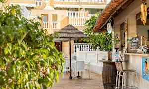 Ulkoterassi, jossa on tuolit ja aurinkovarjo lähellä keltaisen julkisivun ja valkoisten kaiteiden rakennusta. (Tekoälyn luoma teksti)