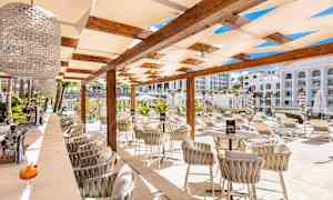 Ulkoilmapaikka, jossa on tuoleja ja pöytiä pergolan alla, ympärillä aurinkotuoleja ja vehreyttä. (Tekoälyn luoma teksti)