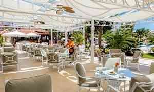 Ulkoterassi, jossa aurinkosuojia, pöytiä ja tuoleja sekä vihreyttä taustalla. (Tekoälyn luoma teksti)