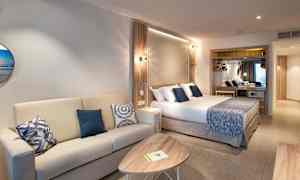 Hotellihuone, jossa on sohva, sohvapöytä ja sinisiä koristeellisia tyynyjä oleva sänky. (Tekoälyn luoma teksti)