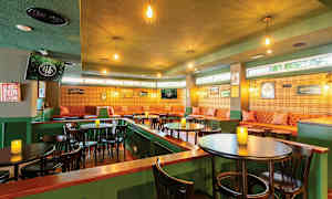 Kodikkaasti sisustettu kahvila tai pubi vihreillä seinillä, riippuvien lamppujen valaistuksessa, ruskeilla pöydillä ja tuoleilla sekä istuma-alueella pehmeillä sohvilla ja seinäkoristeilla. (Tekoälyn luoma teksti)