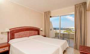 Hotellihuone, jossa on kaksi erillistä sänkyä, puinen pääty ja yöpöydät. Lasipaneelit, jotka tarjoavat näkymät merelle ja parvekekalusteet. (Tekoälyn luoma teksti)