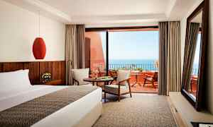 Hotellihuone, jossa sänky, pöytä ja istuinryhmä, terassilta näkymä merelle. (Tekoälyn luoma teksti)