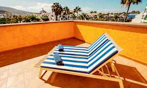 Näkymä aurinkoterassilta, jossa kaksi aurinkotuolia sinivalkoraidallisilla istuinpehmusteilla ja pyyhkeillä, ympäröitynä palmuilla ja rakennuksilla. (Tekoälyn luoma teksti)