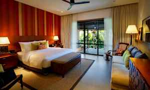 Tilava makuuhuone, jossa on suuri sänky, kaksi yöpöytää lampuilla, sohva, nojatuoli ja parveke, jolta on näkymä vehreään maisemaan. (Tekoälyn luoma teksti)