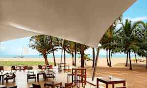 Ulkoravintola valkoisen katoksen alla, näkymä hiekkarannalle, palmuihin ja mereen. (Tekoälyn luoma teksti)