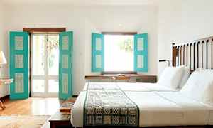 Valoisa makuuhuone, jossa on iso sänky, valkoiset lakanat ja turkoosit ikkunaluukut, jotka päästävät sisään luonnonvaloa. (Tekoälyn luoma teksti)