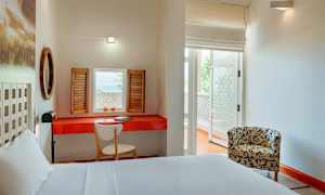 Valoisa hotellihuone, jossa on valkoinen sänky, punainen pöytä ja työtuoli sekä ikkunaluukut ikkunassa. Huoneen toisella puolella on kuvioitu nojatuoli. (Tekoälyn luoma teksti)