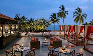 Ulkona sijaitseva ravintola, jossa on pöytiä, tuoleja ja sohvia, palmuja ympärillä ja meri näkyvissä taustalla. (Tekoälyn luoma teksti)