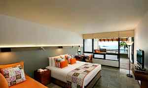 Hotellihuone, jossa on moderni sisustus, sisältäen parivuoteen, värikkäitä tyynyjä ja päiväpeitteen. Huoneessa on myös kirjoituspöytä, sohva ja televisio. Parvekkeelta avautuu näkymä merelle. (Tekoälyn luoma teksti)