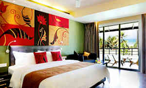 Hotellihuone, jossa on suuri sänky, värikkäät tyynyt, oleskelualue ja parveke, jolta on näkymä hiekkarannalle palmujen ympäröimänä. (Tekoälyn luoma teksti)