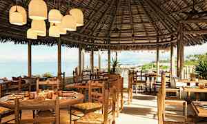 Avoin rantaviivan lähellä oleva ravintola-alue, jossa puiset pöydät ja tuolit sekä olkikatto. (Tekoälyn luoma teksti)