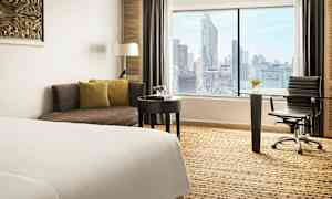 Kuva hotellihuoneesta, jossa on sänky, sohva, kirjoituspöytä ja tuoli. Suuri ikkuna tarjoaa näkymän kaupunkiin. (Tekoälyn luoma teksti)