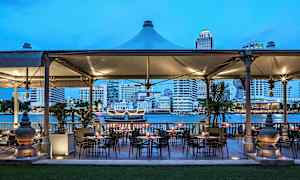 Ulkoterassi, jossa on pöytiä ja tuoleja, sijaitsee joen rannalla, taustalla kaupunki. (Tekoälyn luoma teksti)