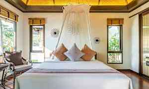 Makuuhuone, jossa on suuri sänky, koristeltu tyynyillä ja valkoinen katos. Huoneessa on ikkunat, joista näkyy vehreyttä. (Tekoälyn luoma teksti)