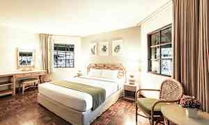 Hotellihuone, jossa on suuri sänky, kaksi lamppua, työpöytä ja tuoli pöydän vieressä, jossa on kukka. (Tekoälyn luoma teksti)