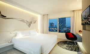 Moderni hotellihuone, jossa iso sänky, ikkuna näkymällä kaupunkiin, tuoli ja televisio. (Tekoälyn luoma teksti)