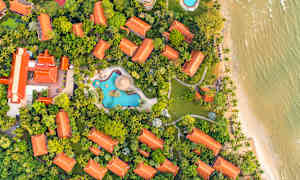 Ilmakuva lomakeskuksesta, jossa on punaiset katot, vehreää kasvillisuutta, uima-altaita ja ranta. (Tekoälyn luoma teksti)