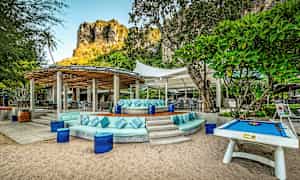 Ulkoterassi, jossa on istuinryhmiä ja pöytiä aurinkosuojakatoksen alla, ympäröitynä vehreydellä ja taustallaan kallio. (Tekoälyn luoma teksti)