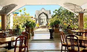 Ulkoterassi, jossa puiset tuolit ja pöydät, ympäröity vehreydellä ja koristeellisilla kasveilla. Taustalla näkyy arkkitehtoninen rakenne. (Tekoälyn luoma teksti)