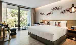 Kuva nykyaikaisesta hotellihuoneesta, jossa on suuri sänky, koristeelliset tyynyt, istuimet ja lasiseinä, joka avautuu terassille ja vehreälle ulkoalueelle. (Tekoälyn luoma teksti)