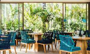 Ravintola, jossa punottuja tuoleja ja puupöytiä trooppisen kasvillisuuden keskellä. (Tekoälyn luoma teksti)