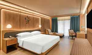 Moderni hotellihuone, jossa on suuri sänky, yöpöydät, nojatuoli, työpöytä ja suuret ikkunat verhoineen. (Tekoälyn luoma teksti)