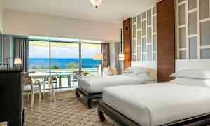 Hotellihuone, jossa on kaksi erillistä yhden hengen vuodetta, modernit kalusteet ja vaalea sisustus. Suuri ikkuna, josta on näkymä merelle. (Tekoälyn luoma teksti)