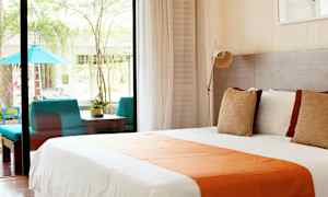 Kuva valoisasta hotellihuoneesta, jossa on suuri sänky, jossa on valkoiset ja oranssit vuodevaatteet, suuret ikkunat avautuvat terassille, jossa on istuinalue ja kasvillisuutta. (Tekoälyn luoma teksti)