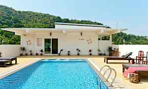 Uima-allas, jossa sinistä vettä, ympäröitynä aurinkotuoleilla ja valkoisella rakennuksella taustalla. Vihreitä kukkuloita näkyy rakennuksen takana. (Tekoälyn luoma teksti)