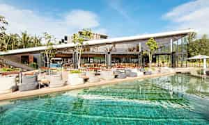 Nykyaikainen kahvila tai ravintola, jossa on suuret lasiseinät, sijaitsee uima-altaan vieressä kirkkaan sinisen taivaan alla. (Tekoälyn luoma teksti)