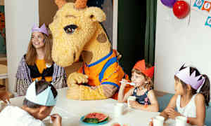 Kirahvimaskotti istuu pöydässä lasten kanssa, joilla on paperikruunut päässään. (Tekoälyn luoma teksti)