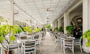 Tämä on ulkoterassi, jossa on valkoisia pöytiä ja tuoleja, ympäröitynä vehreydellä ja varjoisella katoksella. (Tekoälyn luoma teksti)