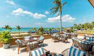 Ulkoterassi, jossa pöytiä ja tuoleja, taustalla meri ja palmuja. (Tekoälyn luoma teksti)