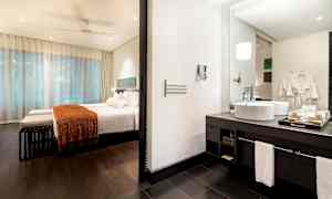 Hotellihuone, jossa on makuuhuone ja kylpyhuone. Makuuhuoneessa on sänky, jossa on valkoinen vuodevaate ja oranssi viltti. Kylpyhuoneessa on pesuallas ja peili. (Tekoälyn luoma teksti)