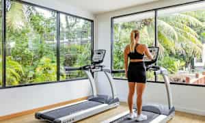 Henkilö katsoo juoksumaton näyttöä kuntosalilla, jossa on suuret ikkunat ja näkymät vehreyteen. (Tekoälyn luoma teksti)