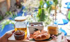 Tarjotin, johon on aseteltu erilaisia leivoksia sekä lasipurkki hunajaa; taustalla näkyy epätarkasti kasveja ja aurinkovarjoja. (Tekoälyn luoma teksti)