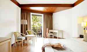 Hotellihuone, jossa on valkoinen työpöytä, sänky ja istuimet sekä parveke, jolta on näkymä yli puiden. (Tekoälyn luoma teksti)