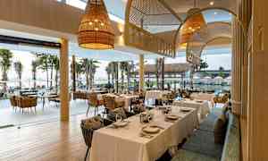 Ravintola, jossa katettuja pöytiä ja näkymä palmujen ja meren yli suurien ikkunoiden kautta. (Tekoälyn luoma teksti)