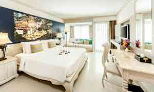 Vaalea ja elegantti hotellihuone, jossa on suuri sänky, oleskelualue ikkunan vieressä ja kirjoituspöytä tuoleineen. (Tekoälyn luoma teksti)