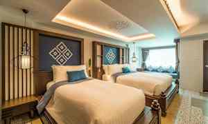 Hotellihuone, jossa on kaksi leveää sänkyä, koristeltu valkoisilla lakanoilla ja sinisillä tyynyillä, sekä suuri ikkuna, jossa on verhot. (Tekoälyn luoma teksti)