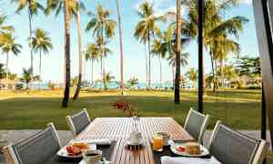 Ulkotila, jossa aamiaista pöydällä. Taustalla nurmikkoa, palmuja ja meri. (Tekoälyn luoma teksti)