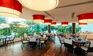 Ravintola, jonka katossa on suuria punaisia lamppuja, ikkunat tarjoavat näkymän vehreille puille ja pöydät ovat katettu. (Tekoälyn luoma teksti)
