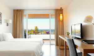 Hotellihuone, jossa on kaksi sänkyä, kirjoituspöytä ja näkymä parvekkeelle, taustalla meri. (Tekoälyn luoma teksti)