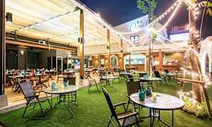 Ulkoilmaravintola, jossa pöytiä ja tuoleja, nurmikko ja valoketjuja. (Tekoälyn luoma teksti)