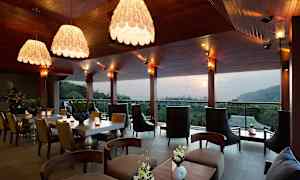 Moderni ravintola tai oleskelutila, jossa on tyylikäs sisustus, suuret ikkunat ja näkymä metsäiseen ja kukkulaiseen maisemaan. (Tekoälyn luoma teksti)