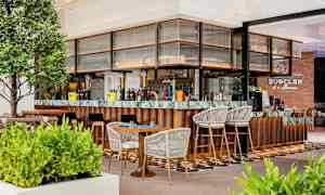 Moderni baari, jossa on puu- ja marmoriyksityiskohtia, ympäröitynä vihreillä kasveilla sekä istumapaikoilta, joissa on tuoleja ja pöytiä. (Tekoälyn luoma teksti)